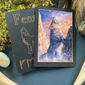 Puede incluir: Dos cartas negras con borde dorado. Una carta presenta una ilustración de lobo aullando con cadenas, la otra tiene la palabra "Fennr" y una silueta de lobo. Las cartas son parte de una baraja, posiblemente cartas de tarot u oráculo.