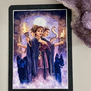 K&ouml;nnte beinhalten: Eine Tarotkarte, die eine dreik&ouml;pfige Frau mit einer Schlange um den Hals zeigt. Sie h&auml;lt Fackeln und einen Schl&uuml;ssel. Zwei schwarze Hunde stehen an ihrer Seite. Die Karte ist vor einem dunklen Hintergrund mit einem Mond am Himmel platziert.