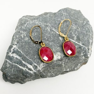 May include: A pair of gold-colored leverback earrings with oval-shaped, faceted, deep red gemstones. The earrings are set in a gold-colored bezel and rest on a gray stone surface against a white background. The gemstones have a brilliant cut.
