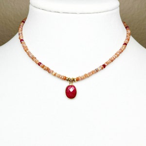 May include: A delicate gold necklace with a red gemstone pendant. The necklace is made of small, light brown beads.