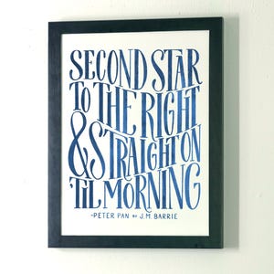 May include: A framed wall print featuring the blue text Second Star To The Right & Straight On Til Morning - Peter Pan by J.M. Barrie. The artwork is displayed in a simple black frame with a white background and bold, stylized blue lettering.