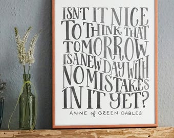 May include: A rectangular framed print with a white background and dark gray hand-lettered text that reads: ISN'T IT NICE TO THINK THAT TOMORROW IS A NEW DAY WITH NO MISTAKES IN IT YET? ANNE OF GREEN GABLES. The frame is a thin, light brown wood.