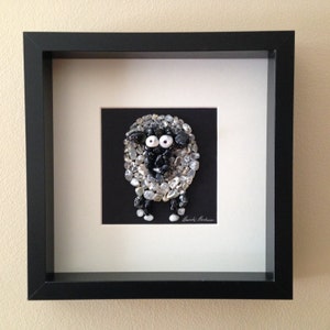 May include: A framed artwork featuring a sheep made of small, gray and black stones. The sheep has large white eyes and is set against a black background. The artwork is in a black frame with a white mat.