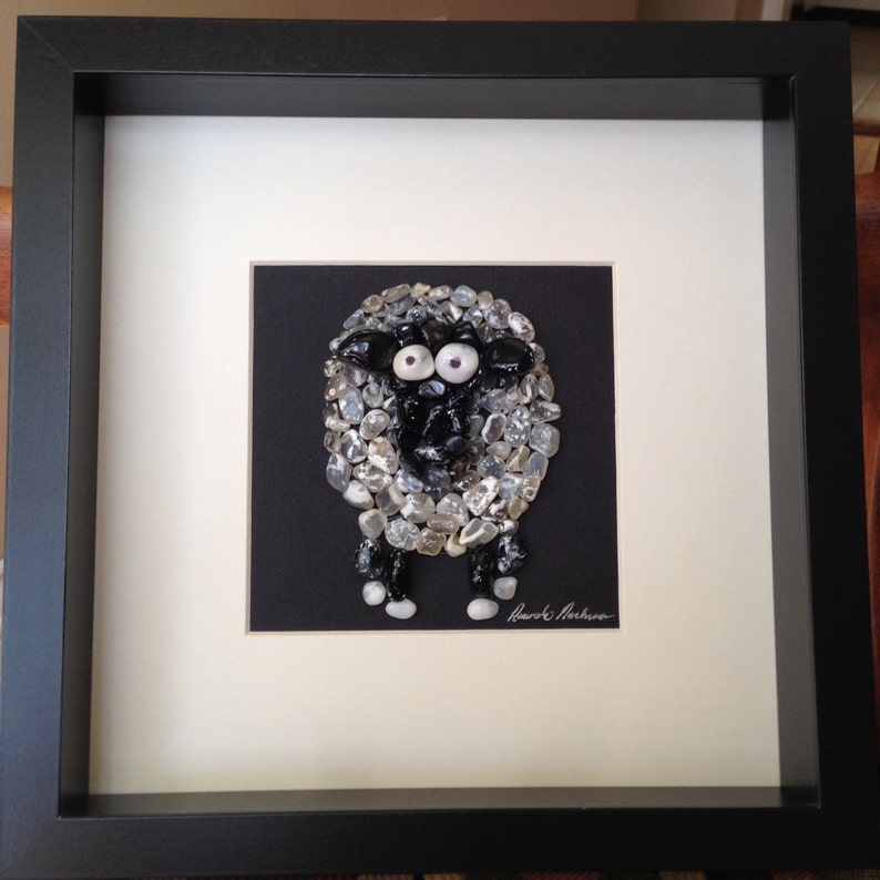 May include: Framed artwork featuring a sheep made of small, light-colored stones on a black background. The sheep has a black face and legs, with large, googly eyes. The artwork is set in a black frame with a white mat.