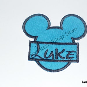 Peut inclure: Un écusson brodé bleu vif en forme de silhouette de tête de souris avec une bordure bleu foncé. Une bannière rectangulaire bleu foncé est cousue au centre, avec le nom LUKE brodé en fil noir dans une police de caractères stylisée.