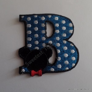 May include: An embroidered letter B patch featuring a blue background with white polka dots and a black outline. A black silhouette of a mouse head with a small red bow tie is positioned over the lower left section of the letter B.