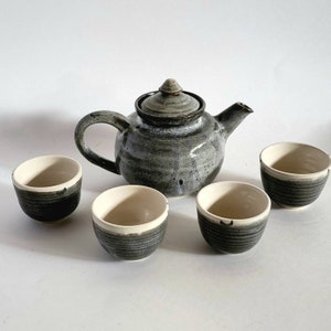 May include: A set of four ceramic teacups and a teapot with a speckled gray and white glaze. The teapot has a rounded body and a small lid with a knob. The teacups are all the same size and have a white interior and a gray exterior.