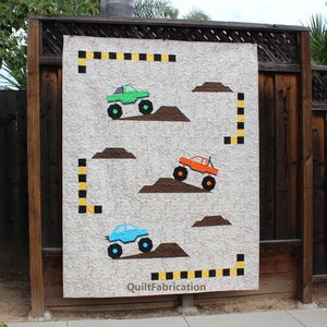 Pode incluir: Um edredom branco com um design de caminhão monstro marrom, laranja, verde e azul. O edredom tem uma borda quadriculada preta e amarela e o texto "QuiltFabrication" no canto inferior direito.