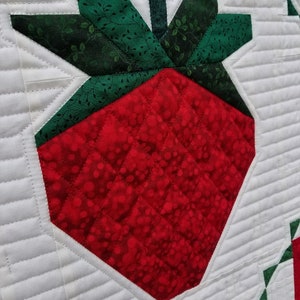 May include: A close-up of a red and green quilted strawberry with a white background. The strawberry is made of fabric and has a quilted design. The text "QuiltFabrication" is visible in the bottom right corner of the image.