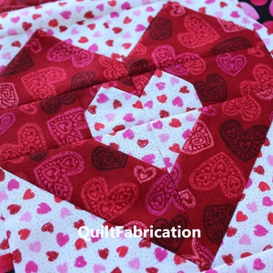 Puede incluir: Un bloque de colcha rojo y blanco con una gran forma de corazón en el centro. El corazón está hecho de tela roja con un pequeño patrón de corazón blanco. La tela de fondo es blanca con un pequeño patrón de corazón rosa. El texto "QuiltFabrication" es visible en el centro inferior de la imagen.