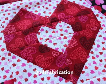 May include: A red and white quilt block with a large heart shape in the center. The heart is made of red fabric with a small white heart pattern. The background fabric is white with a small pink heart pattern. The text "QuiltFabrication" is visible in the lower center of the image.