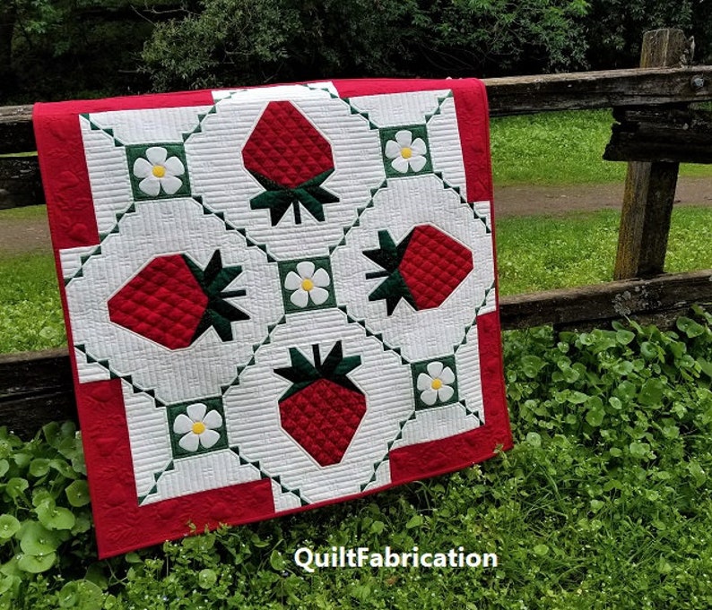 May include: A red, white, and green quilt with a strawberry and flower pattern. The quilt is hanging on a wooden fence. The text "QuiltFabrication" is visible in the bottom right corner.