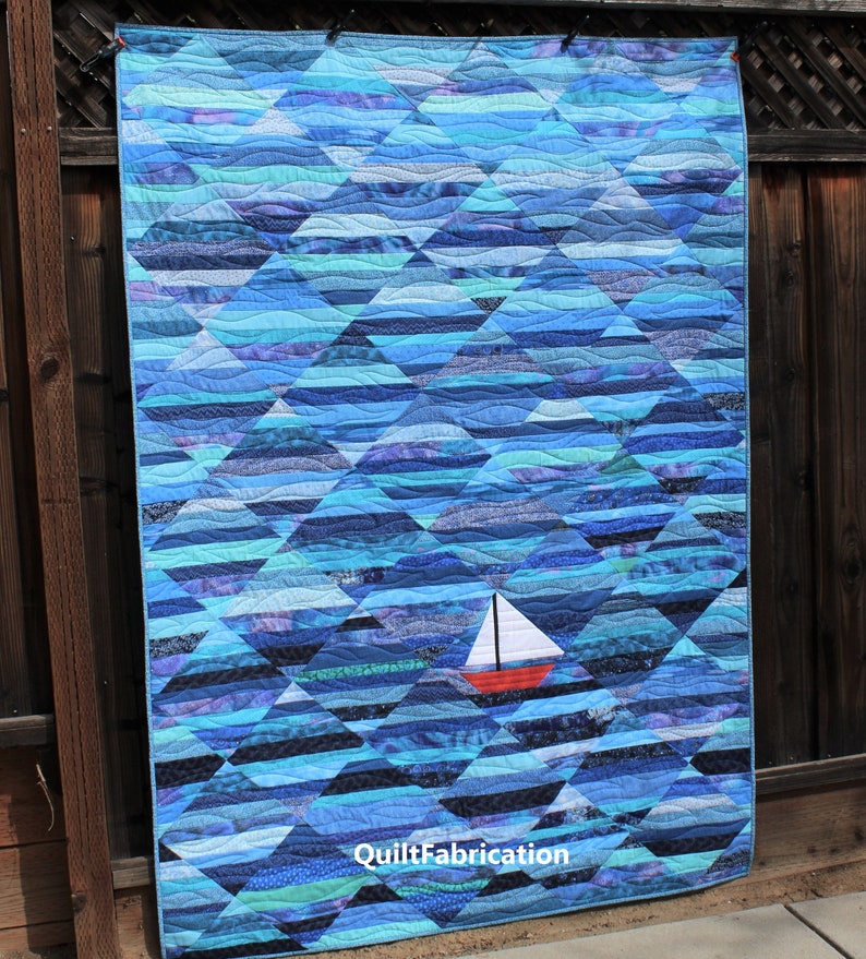 May include: A blue and white quilt with a sailboat design. The quilt is made of many different shades of blue and white fabric, and the sailboat is a bright red with a white sail. The quilt is hanging on a wooden fence. The text "QuiltFabrication" is visible in the bottom right corner of the quilt.