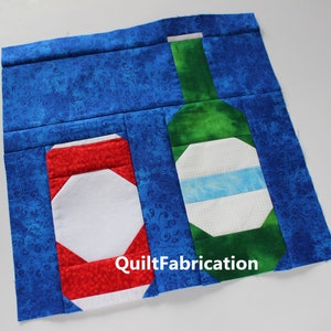 May include: A quilt block with a red, white, and blue can and a green and white bottle on a blue background. The text "QuiltFabrication" is visible in the lower right corner.