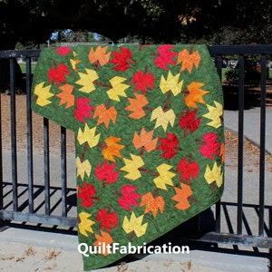 May include: A green quilt with a pattern of yellow, orange, and red maple leaves. The quilt is hanging on a black metal fence. The text "QuiltFabrication" is visible in the bottom right corner of the image.