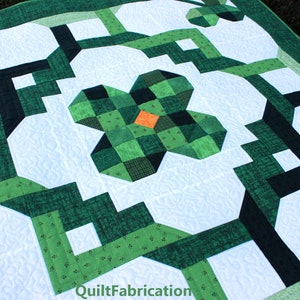 May include: A handmade quilt with a shamrock design. The quilt features a white background with green and black geometric shapes. The shamrock in the center has an orange square in the middle. The quilt is made of fabric and has a quilted texture.
