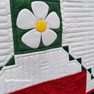 May include: A white quilt with a green and white flower applique. The quilt has a red border with a quilted design. The text "QuiltFabrication" is visible in the lower left corner.