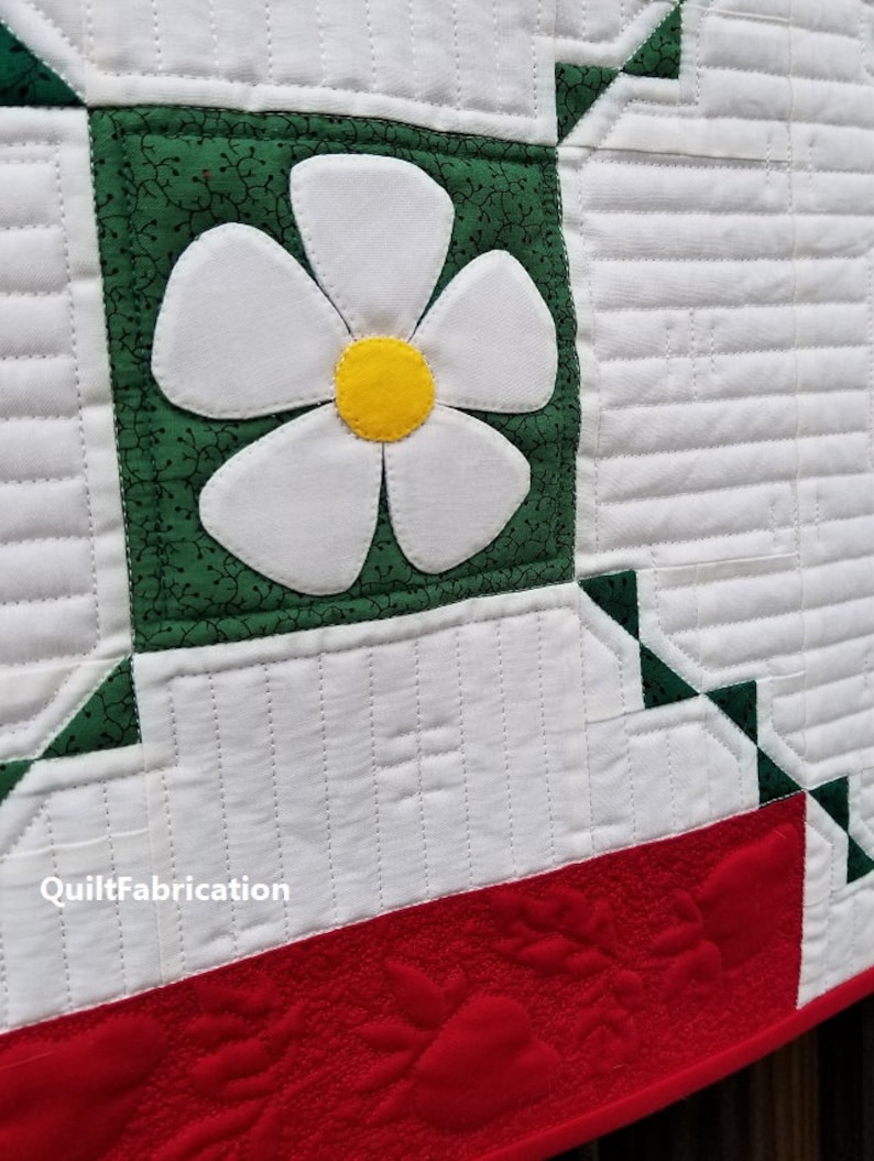 May include: A white quilt with a green and white flower applique. The quilt has a red border with a quilted design. The text "QuiltFabrication" is visible in the lower left corner.