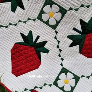 May include: A red and green patchwork quilt with a strawberry and white flower design. The quilt is made with different fabrics and has a quilted pattern. The text "QuiltFabrication" is visible in the lower right corner.