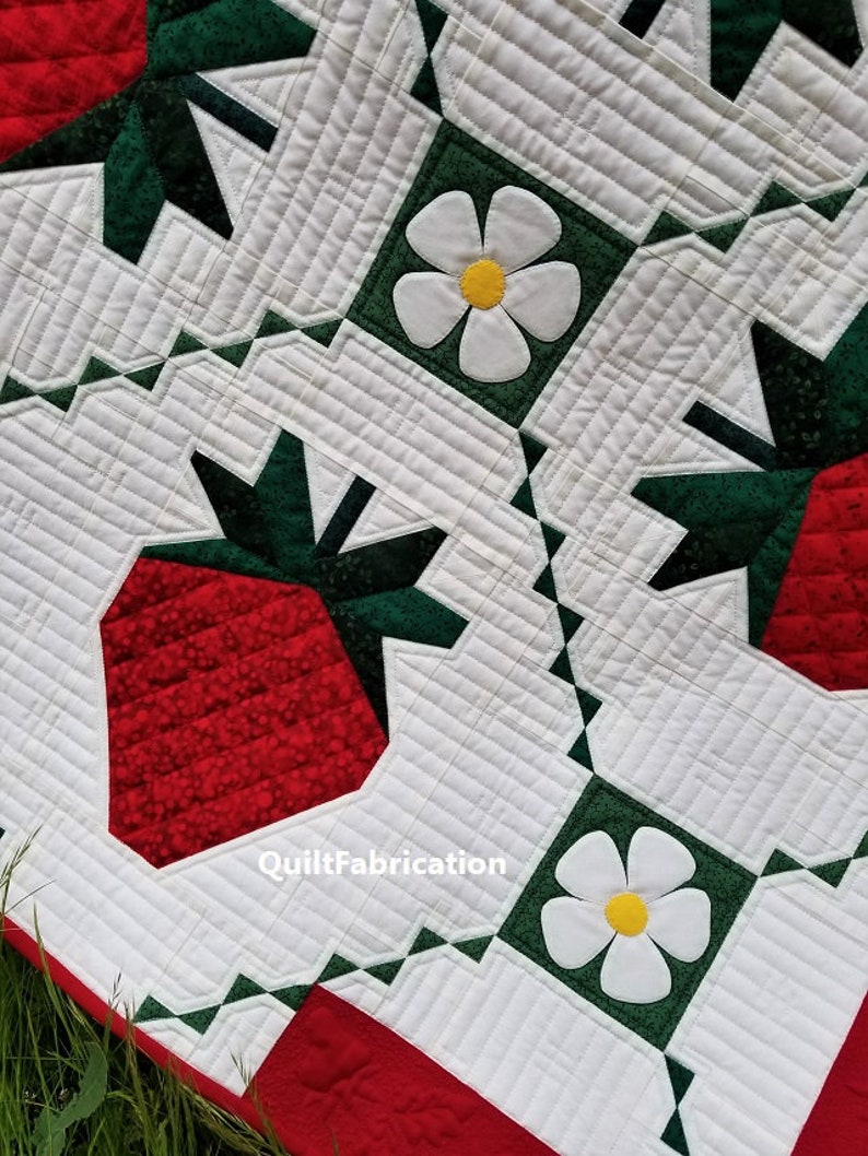 May include: A red and green patchwork quilt with a strawberry and white flower design. The quilt is made with different fabrics and has a quilted pattern. The text "QuiltFabrication" is visible in the lower right corner.