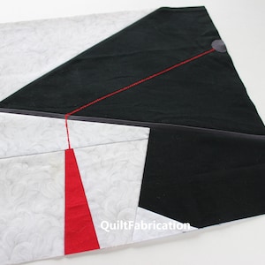 Puede incluir: Un bloque de colcha con un diseño de tela negra, roja y blanca. El bloque está cosido con hilo rojo. El texto "QuiltFabrication" es visible en la esquina inferior derecha.