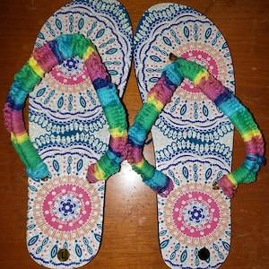 May include: A pair of silver flip flops with a colorful, multi-colored braided strap. The flip flops have a mandala design with pink, blue, and orange accents.