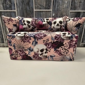May include: A pink and black tissue box cover with a floral and skull design. The cover has a couch shape with four pillows, each with a skull design.