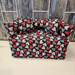 Puede incluir: Una funda para caja de pañuelos de color negro con un patrón de corazones rojos y blancos. La funda tiene cuatro almohadas con el mismo patrón de corazones.