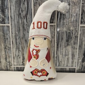 May include: A whimsical white gnome doll with a long, curved hat embroidered with the number "100" in red. The doll has blonde hair, a smiling face, and a white dress with red accents and heart-shaped designs. A silver pom-pom adorns the hat.