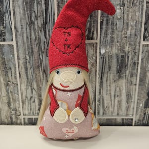 May include: A handmade gnome doll with a red, glittery hat and long blonde hair. The hat features a heart with "TS + TK" embroidered on it. The doll wears a pink dress with the text "TS + TK FOREVER" and a red jacket. A decorative item.