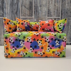 May include: A colourful tissue box cover with a couch design. The cover features a tie-dye pattern with paw prints and cartoon dogs. The couch has four cushions with the same pattern.