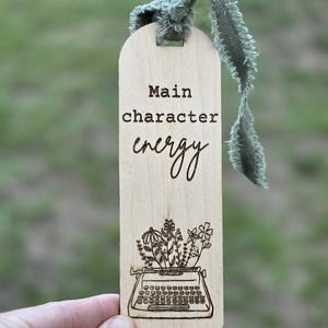 May include: Wooden bookmark with the text "Main character energy" and a drawing of a typewriter with flowers. The bookmark has a green ribbon attached at the top.