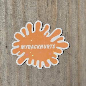 May include: A sticker with an orange splatter design and the text "MYBACKHURTS" in white. The sticker has a white border and small star accents. The sticker is on a wooden surface.