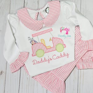 May include: A white long-sleeved shirt with a pink gingham collar and cuffs. The shirt features an embroidered design of a golf cart with the text "Daddy's Caddy".