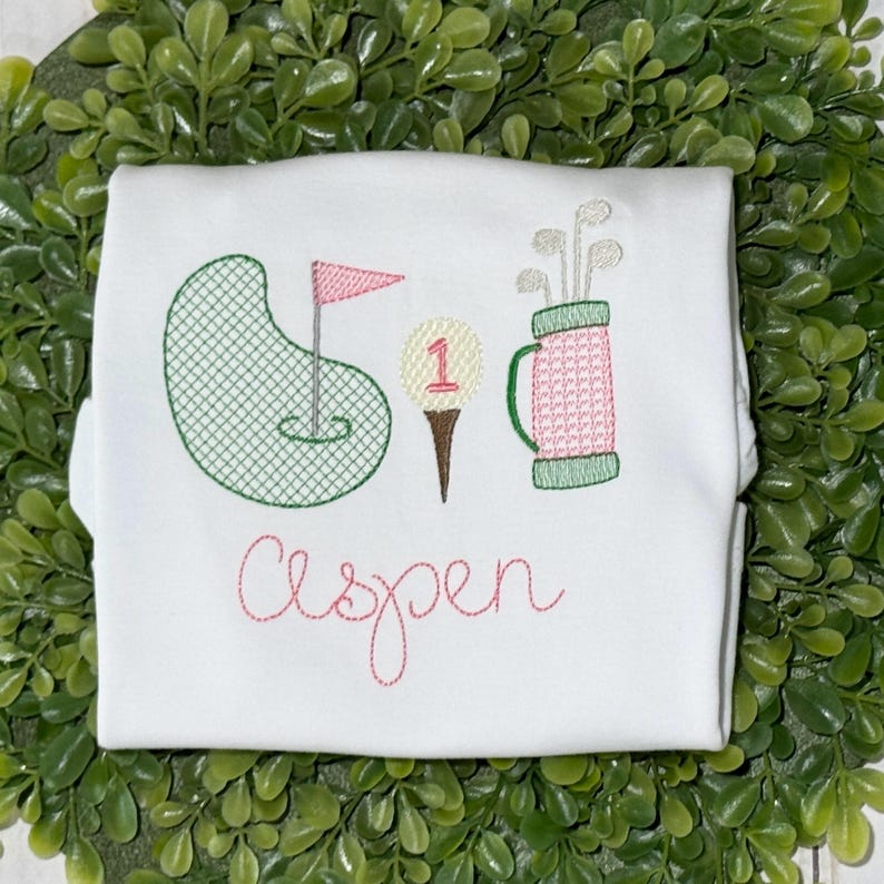 May include: White onesie with embroidered green golf course, pink flag, brown golf club, white golf ball with a red "1", and a pink and green golf bag with clubs. The onesie also has the name "Aspen" embroidered in pink.