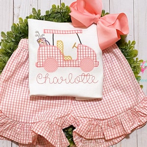 May include: A pink and white gingham outfit with a golf cart design and the name "Charlotte" embroidered on the front. The outfit includes a short-sleeved shirt and a pair of shorts with a ruffled hem.
