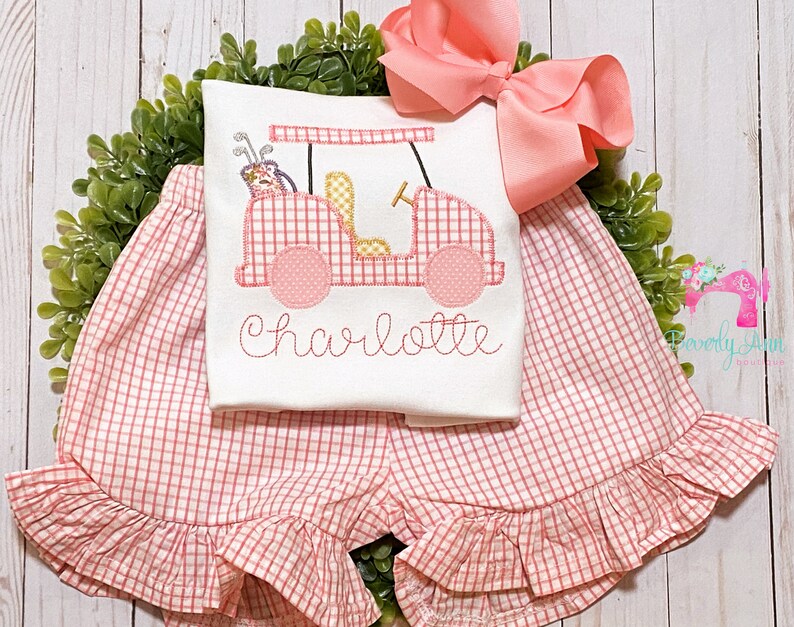 May include: A pink and white gingham outfit with a golf cart design and the name "Charlotte" embroidered on the front. The outfit includes a short-sleeved shirt and a pair of shorts with a ruffled hem.