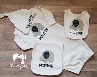 May include: A white baby outfit set with an elephant design and the name "Brayden" embroidered on each piece. The set includes a hat, a long-sleeved onesie, a bib, and a pair of pants.