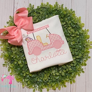 May include: White onesie with a pink and white gingham golf cart design and the name "Charlotte" embroidered in pink.