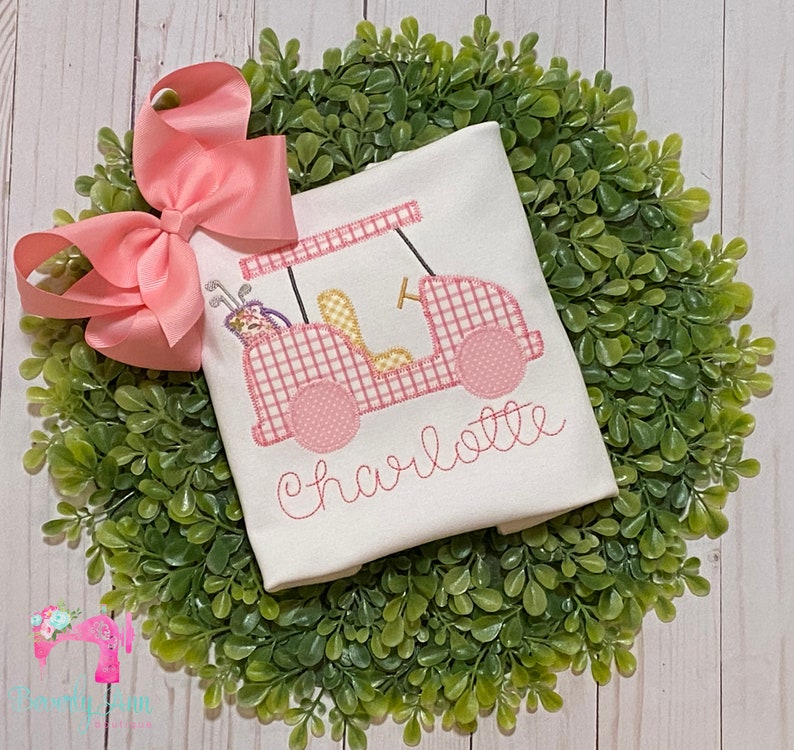 May include: White onesie with a pink and white gingham golf cart design and the name "Charlotte" embroidered in pink.