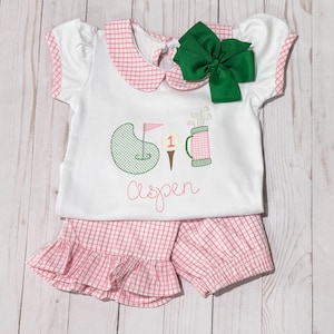 May include: A pink and white gingham baby outfit with a green embroidered golf design and the name "Aspen".