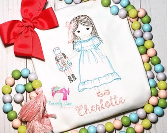 May include: A white embroidered shirt with a girl holding a nutcracker doll. The girl has brown hair and is wearing a blue dress. The shirt has the name "Charlotte" embroidered on it.
