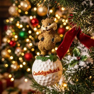 May include: A hanging Christmas tree ornament featuring a brown gopher figure standing atop a white golf ball. The golf ball has the word Caddyshack printed in red lettering. The ornament includes a silver metal hook for hanging on a branch.