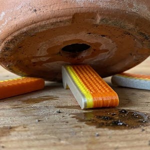 May include: Three L-shaped pot feet with a textured orange top surface and a light gray base. These wedge-shaped risers are positioned underneath a terracotta plant pot to elevate it, creating a small gap between the pot and the surface.