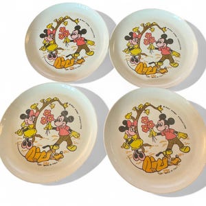 May include: A set of four round, off-white plates featuring a colorful graphic of Mickey Mouse, Minnie Mouse, and Pluto. The illustration shows Mickey holding red flowers for Minnie under a tree, with Pluto lying nearby. Text reads Walt Disney Productions.