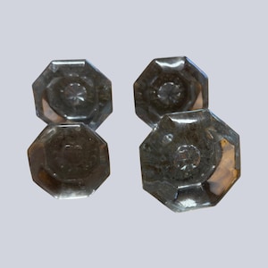 May include: A set of four clear glass door knobs, each featuring an octagonal shape with a faceted surface and a central starburst pattern. The knobs are transparent with subtle light reflections visible on the edges of the glass.