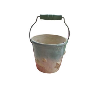 May include: A ceramic planter shaped like a bucket with a green handle. The planter has a light blue and white glaze with a seashell design.