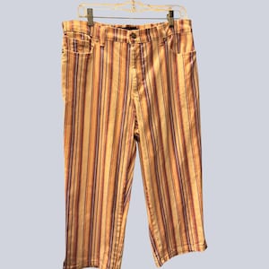 May include: Mid-length pants featuring a vertical striped pattern in shades of yellow, orange, and purple. These trousers include a standard waistband with a button closure, belt loops, and two front pockets, hanging from a wire clothing hanger.