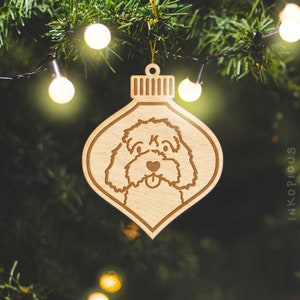 May include: A wooden Christmas ornament shaped like a bauble with a dog's face carved into it. The dog has a tongue sticking out and a heart-shaped nose.
