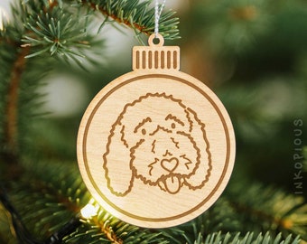 May include: A wooden Christmas ornament shaped like a bauble with a cartoon doodle of a dog's face. The dog has a heart on its nose and its tongue is sticking out.